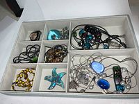 Top view of divided jewelry box containing multiple Hawaiian blown glass jewelry pieces including pendants, necklaces, and beads in various colors and styles.