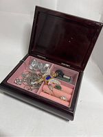 Open dark reddish wood jewelry box showing pink velvet compartments holding assorted jewelry pieces including a Tinker Bell pendant