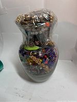 Front view of the glass jar fully packed with assorted jewelry pieces visible through the clear glass