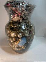 Glass jar filled with various costume jewelry pieces showing metallic earrings, brooches, pearls, and beads visible through clear glass
