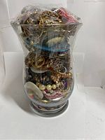 Front view of the large glass jar packed with various jewelry pieces visible through the top plastic wrapping.