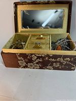 Mini jewelry box open showing brown and white flowered fabric exterior, internal mirror, and compartments filled with assorted rings and jewelry.