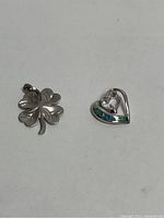 Photo showing both pendants: a four leaf clover pendant and a heart-shaped pendant with opal inlay.