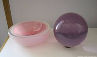 Pink glass bowl and lavender glass sphere displayed side by side on white surface