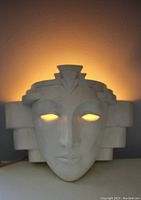 Front view of ceramic face sconce lit from behind, showing eye cutouts and geometric details