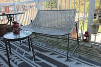 White plastic rattan loveseat with metal frame alongside round glass top table and two red metal candle lanterns on a geometric patterned outdoor rug on wooden decking.