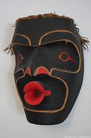 Front view of black painted cedar mask with red projecting lips, stylized eyes and facial design, cedar bark trim visible