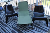 One green Coleman folding patio lounger positioned between two black molded plastic outdoor chairs on a patterned outdoor rug