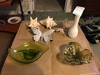 Green and clear art glass bowl, green divided leaf shape bowl, white tall milk glass vase, two butterfly clips, and three sea shells arranged on cardboard.