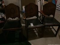 Three wood framed dining chairs with black vinyl seats including one with wooden arms, showing some wear and scuffs on seats. Chairs have carved backrests with an oval motif.