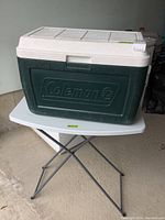 Green Coleman cooler with white lid on top of small white folding table with metal legs