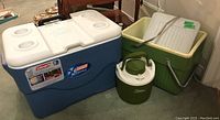 Photo showing the blue Coleman Xtreme cooler, green Coleman drink jug, and pale green/cream Thermos cooler with metal handles and wire rack inside, placed on carpet near furniture.