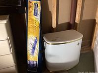 Photo showing the folded camp cot in its blue and yellow storage bag with Broadstone logo, standing upright next to the metal laundry basket.
