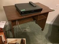Small particleboard desk with wood grain finish, rectangular top surface, and two side panels. A remote and electronic device are placed on top (not included). Visible caster wheels at the bottom for mobility.