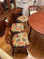 Six wooden dining chairs arranged around a wooden table, with three floral patterned cushions on chairs and some cushions stacked nearby.