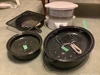Black enamel roaster pans (one round, one oval), cast iron fry pans and grill rack, Black & Decker steamer in background on carpeted floor