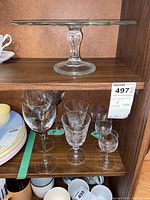 Photo of glass cake stand on upper shelf and 7 wine glasses on lower shelf, some clear crystal with faceted bowls and some with colored spiral stems.