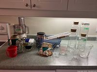 Photo showing overall arrangement of kitchen items including food processor, glass canisters with cork lids, drinkware, a wire basket, and adhesive shelf liner on countertop.
