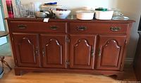 Front view of the Krug Saugeen maple sideboard showing 2 drawers on top and 4 paneled cabinet doors below. Sideboard has polished wood with antique brass hardware. Various dishes placed on top.