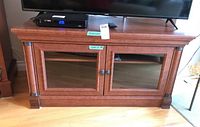 Front view of the wooden media cabinet showing two glass front doors with round knobs, the top surface supporting TV and devices, and brown finish with decorative molding.