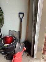 Photo of garden tools including a shovel with a wooden handle and blue D-shaped grip, fan rake with wooden handle, several resin flowerpots in red and black (including a large urn-shaped one), an orange plastic watering can, and a garden hose mounted to the wall.