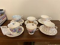 Front view showing all five teacups with their matching saucers on a wooden surface, plus one extra teacup without a saucer, displaying varied floral and geometric patterns.