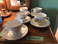 Five vintage teacups paired with their matching saucers displayed on a wooden surface, showing variety of floral designs and gold trims.