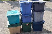 Stack of 8 plastic storage totes in various colors including dark blue, light blue, beige, green, and turquoise, all with lids, showing used condition outdoors in sunlight.