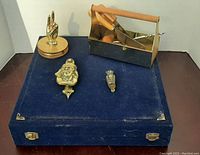 Photo showing entire lot on a blue velvet case surface including magnifying glass, brass knockers, cheese tool set, and decorative plate.