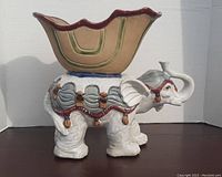 Front angled view of the white ceramic elephant planter showing the detailed saddle and open bowl with scalloped brown and green edge.