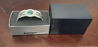Silver cuff bracelet displayed inside original silver-colored insert box with black foam padding, outer dark blue box closed nearby.