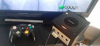 Black Nintendo GameCube console with controller and part of TV screen showing game interface.