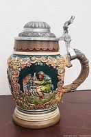 Side view of the ceramic beer stein showing relief artwork of a man stoking a fire, ornate decorative border, and hinged metal lid with detailed embossing.