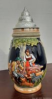 Front view showing detailed scene with man, woman, animals under trees on a glossy black background, with the pewter lid closed.