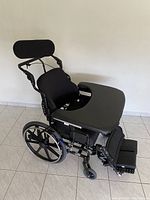 Side and angled views of manual tilt wheelchair showing padded seat, backrest, footrests, headrest, and tray table with black metal frame and wheels.