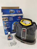 Bissell SpotClean ProHeat cleaner beside unopened Bissell cleaning solution bottle and hose clips with original product box behind.