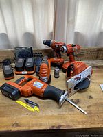 Full view of several Black And Decker Firestorm power tools arranged on wooden bench, showing cordless drill, Sawall, orbital sander, circular saw, battery packs, and charger.
