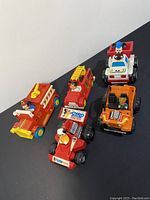 All five vintage Mickey Mouse themed toy vehicles arranged on a black surface showing red fire engine, racing cars, white siren car, and orange buggy.
