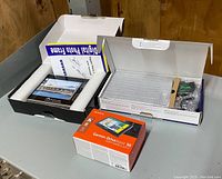 Three boxed items on a gray table against wood wall: Garmin Drive Assist 50 retail box in orange and white, Sony S-Frame digital photo frame box showing a displayed photo, and an additional digital photo frame in black protective packaging with user manual visible.