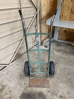 Front angle view of the teal metal hand truck dolly showing two large wheels, metal frame and loading plate with visible rust and wear.