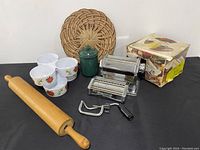Full view of all lot items on black surface: large wooden rolling pin, metal manual pasta roller with crank and cutter, three floral bowls, green jar with lid, woven rattan warmer, and product box