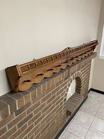 Full view of the wooden wall shelf resting on a brick ledge showing the scalloped detailing and peg hooks.