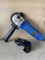 Blue Mastercraft corded electric angle grinder with auxiliary handle attached, placed vertically against wall, with black removable blade guard lying next to it.