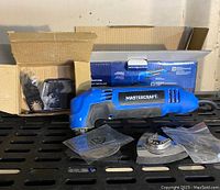 Blue and black Mastercraft oscillating multi tool with several accessories and open cardboard box behind it.