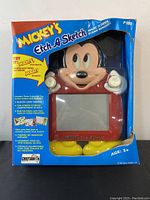 Front of the boxed Mickey's Etch A Sketch featuring Mickey Mouse face and yellow shoes visible through clear plastic with colorful blue box.