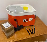 Orange and white electric pottery wheel with silver wheel head, yellow bowl, and trimming tools