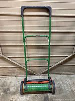 Front view of the green metal push reel lawn mower with black handle and wheels and orange blades visible below.