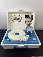 Open record player showing Mickey Mouse graphic inside lid, blue turntable, white platter, and Mickey glove tonearm.