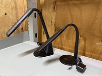 Two black desk lamps on a white table, one identified as OttLite brand with flexible gooseneck and circular base, second has segmented adjustable arm and rectangular light panel.