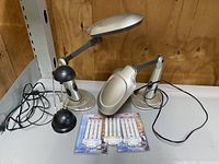 Photo of two silver adjustable Sun Lamps with articulated arms and black bases, alongside two sets of sealed replacement bulbs in packaging.